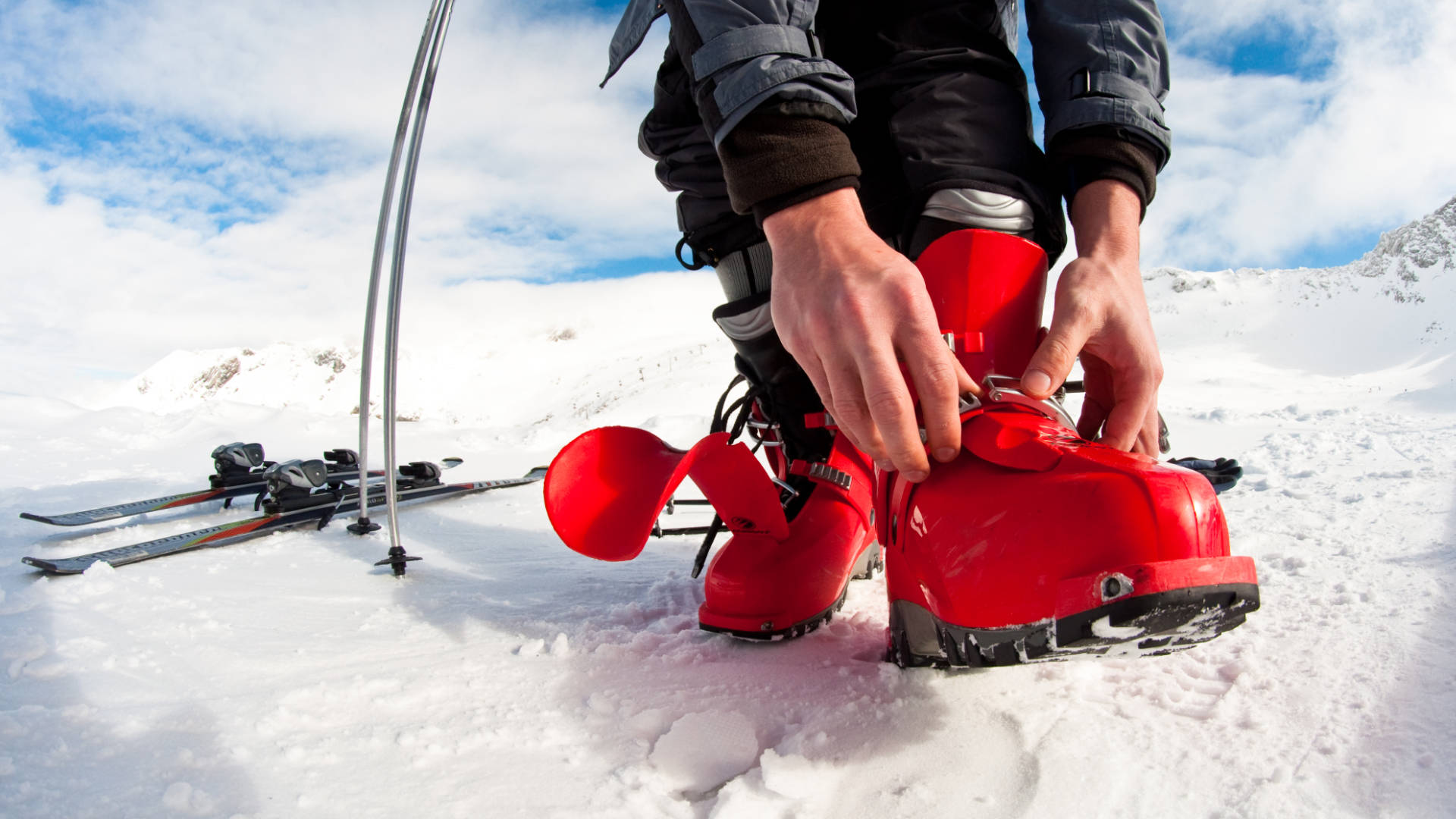 Hands closing the buckles of a red ski boot, with the skis on the snow