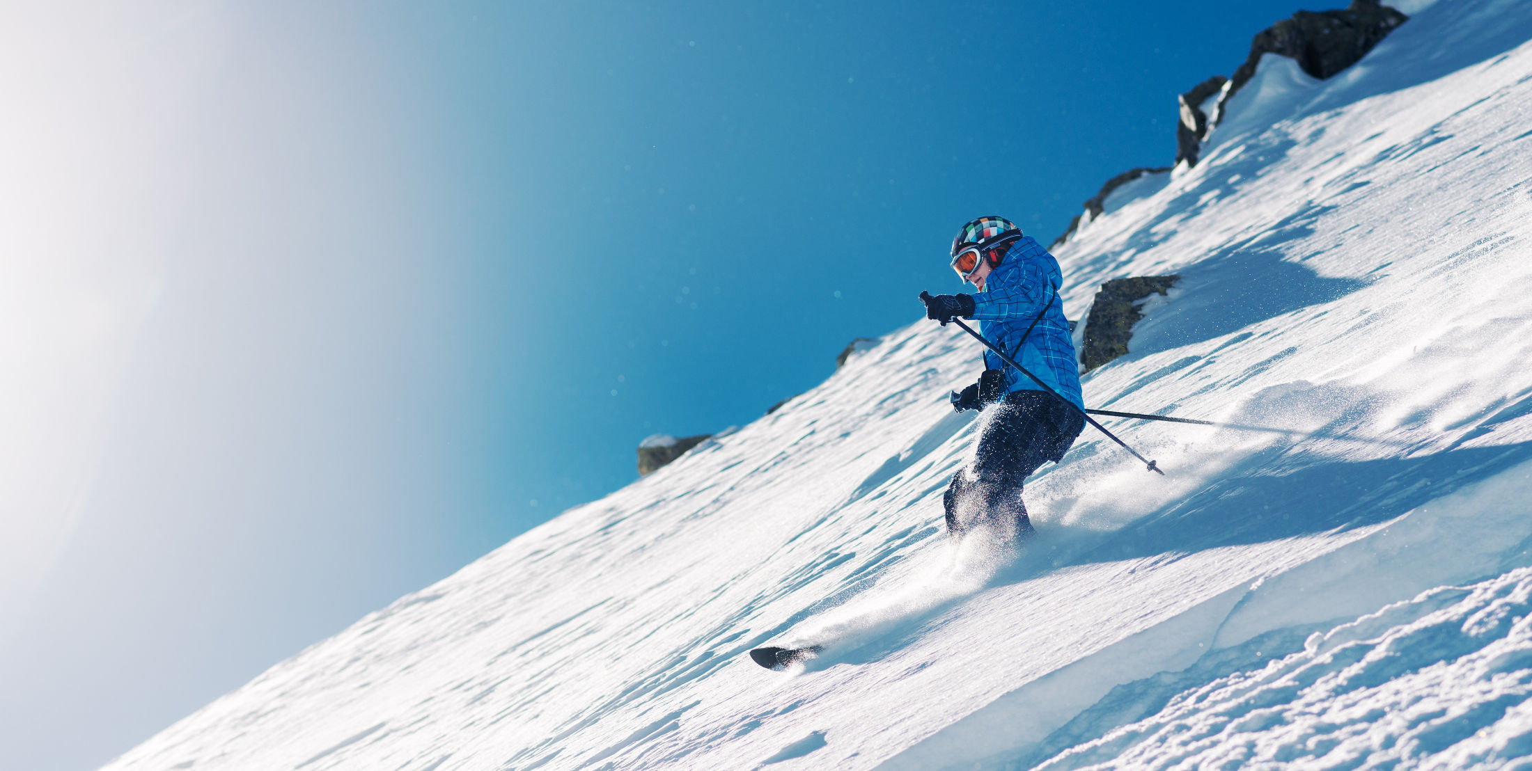 Skier in a blue jacket skiing down a powder slope, blue sky